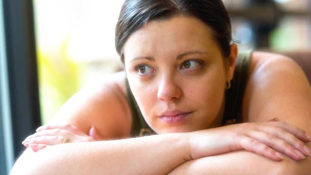 A brunette woman sits with her chin resting on her folder arms, on the table in front of her.